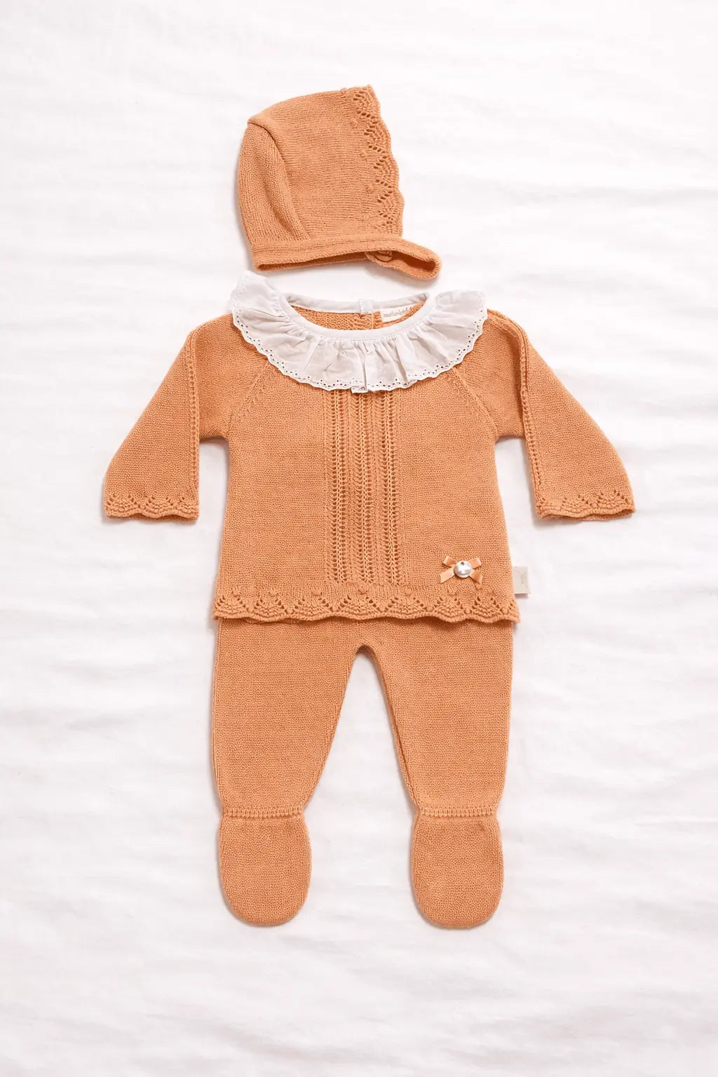 Conjunto de bebé em malha cor salmão com camisola de gola branca em folho, calça com pé e touca combinada, apresentado em fundo branco suave.