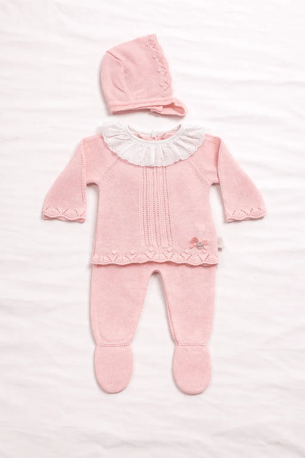 Conjunto de bebé em malha rosa composto por camisola com gola branca em folho, calça com pé e touca combinada sobre fundo branco minimalista.