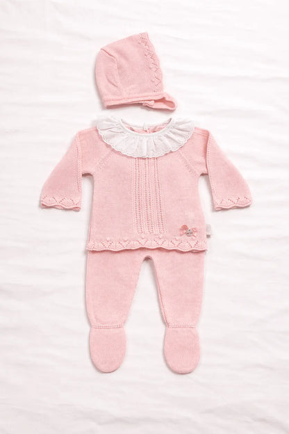 Conjunto de bebé em malha rosa composto por camisola com gola branca em folho, calça com pé e touca combinada sobre fundo branco minimalista.